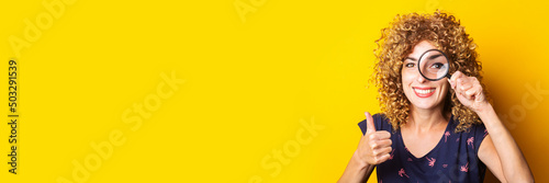 curly young woman looking at camera through magnifying glass showing thumbs up gesture on yellow background. Banner.