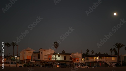 Palm trees silhouettes and full moon in twilight sky, California beach, USA. Beachfront houses or homes on coast in evening, fullmoon on pacific ocean shore in dusk. Lifeguard tower. Light in windows.