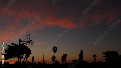 People playing basket ball game  silhouettes of players on basketball court outdoor  sunset ocean beach  California coast  Mission beach  USA. Black hoop  net and backboard on streetball sport field.