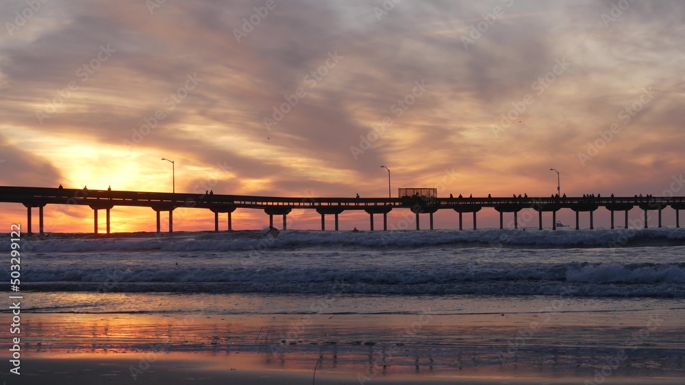 Obraz premium Silhouette of people walking, surfers surfing by pier in sea water. Ocean waves, dramatic sky at sunset. California coast, beach or shore vibes at sundown. Summer seascape seamless looped cinemagraph.