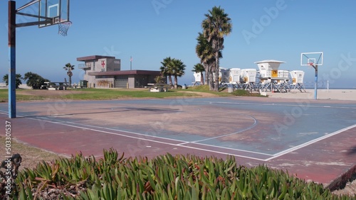 Palm trees and basketball sport field or court on beach, California coast, USA. Streetball playground on shore and lifeguard stand, tower ot station. Mission beach, San Diego. Hoop, backboard and sky.