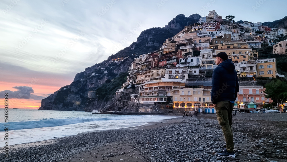 Tourist man watching sunset on Marina Grande Beach and colorful buildings of hillside village Positano at Amalfi Coast, Italy, Campania, Europe. Luxury vacation at Tyrrhenian, Mediterranean Sea
