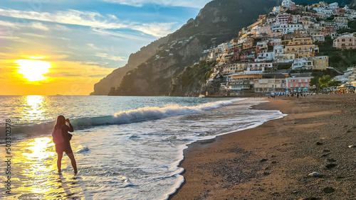 Woman standing in water and watching sunset on Marina Grande Beach and colorful buildings of hillside village Positano, Amalfi Coast, Italy, Campania, Europe. Vacation at Tyrrhenian, Mediterranean Sea photo