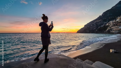 Fototapeta Naklejka Na Ścianę i Meble -  Woman taking pictures while watching sunset on Marina Grande Beach and colorful buildings of hillside village Positano, Amalfi Coast, Italy, Campania, Europe. Vacation at Tyrrhenian, Mediterranean Sea