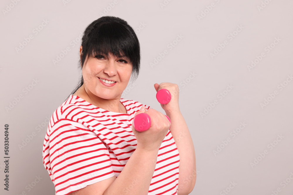 Happy overweight mature woman doing exercise with dumbbells on grey background. Space for text Happy overweight mature woman doing exercise with dumbbells on grey background. Space for text