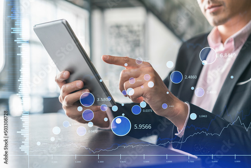 Businessman in suit using tablet device to optimize trading strategy at corporate finance fund. Forex chart hologram over modern panoramic office background