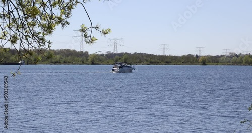 Boat sailing on calm water on the Oude Maas river, green trees and metal power poles in the background, Molenplas nature reserve, sunny day in Stevensweert, South Limburg, Netherlands photo