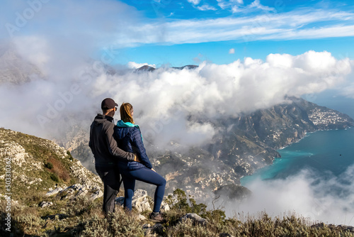 Happy couple standing at cliff with scenic view from Monte Comune on the coastal town Positano. Magical hiking above clouds in Lattari Mountains, Apennines, Amalfi Coast, Campania, Italy, Europe. Awe
