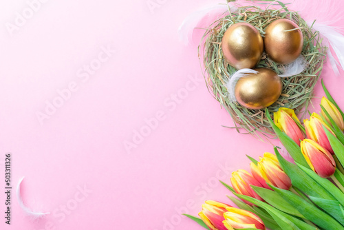 Easter Golden eggs in basket with spring tulips, white feathers on pastel pink background in Happy Easter decoration. Spring holiday flat lay concept.