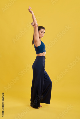 Side view of slim girl standing, raising hand, looking down, with closed eyes. Brunette with pony tail stretching, meditating, yoga pose practicing. Concept of new age and yoga.