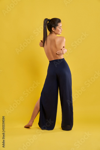 Back view of slim girl with bare back standing backwatds, holding by hands. Female with pony tail yoga doing, looking down, meditating. Isolated on yellow studio background.