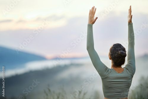 Back view of woman female in green sportswear doing virabhadrasana on blur nature background. Crop of young pretty female standing in warrior pose outdoors in cloudy mountains. Concept of yoga.