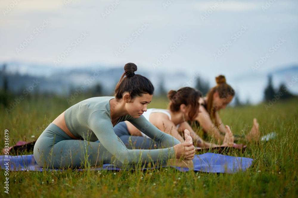 Side view of asian brunette female in green wear stretching leg on grass near blur girls in mountains. Women in group working out practising yoga on foggy nature. Concept of healthy lifestyle. Side view of asian brunette female in green wear stretching leg on grass near blur girls in mountains. Women in group working out practising yoga on foggy nature. Concept of healthy lifestyle.