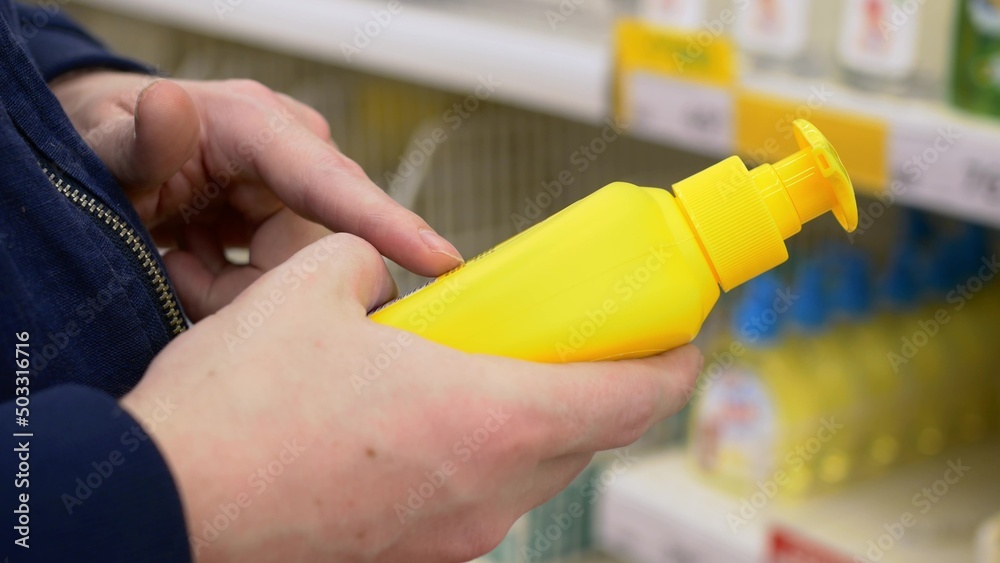Close-up of a yellow plastic bottle with a dispenser, liquid soap in the hands of a buyer who carefully reads the composition. Buying hygiene products in the supermarket.