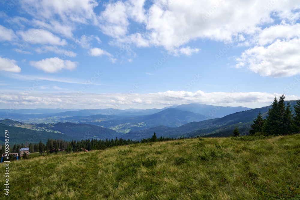 Remote mountain village, with few small houses in coniferous forest in autumn. Panoramic view of magnificent landscape, with rolling hills and cloudy blue sky on background. Concept of highland.