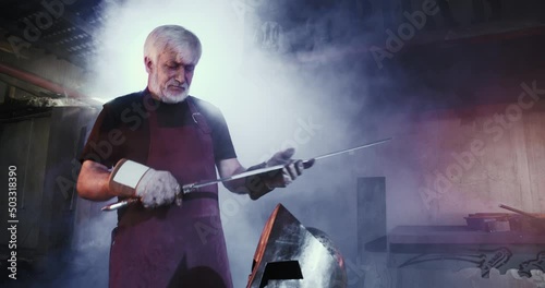 Front view of old blacksmith forging armor for knight, warrior. Male with grey hair and beard holding sword, inspecting, mask on table, smoke spreading. Concept of forging.