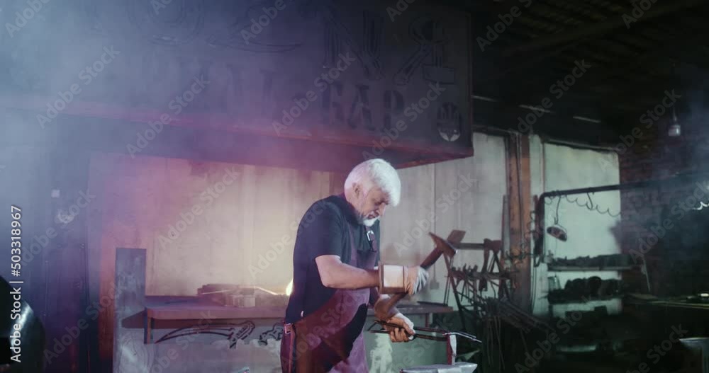 Side view of old blacksmith with grey hair and beard beating with hammer. Man with grey hair and beard wearing apron working in smithy, forging. Concept of heavy craftmanship. Side view of old blacksmith with grey hair and beard beating with hammer. Man with grey hair and beard wearing apron working in smithy, forging. Concept of heavy craftmanship.