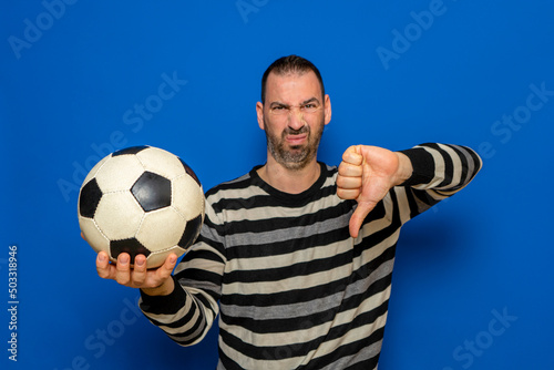 Handsome latin man holding football with angry face, negative sign showing dislike with thumbs down, rejection concept. Isolated on blue background.