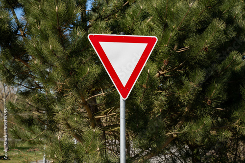 Give way road sign next to crossroads. Also called yield sign to guide traffic. Triangular shape with a red stroke. Warning symbol to beware of the traffic crossing the way.
