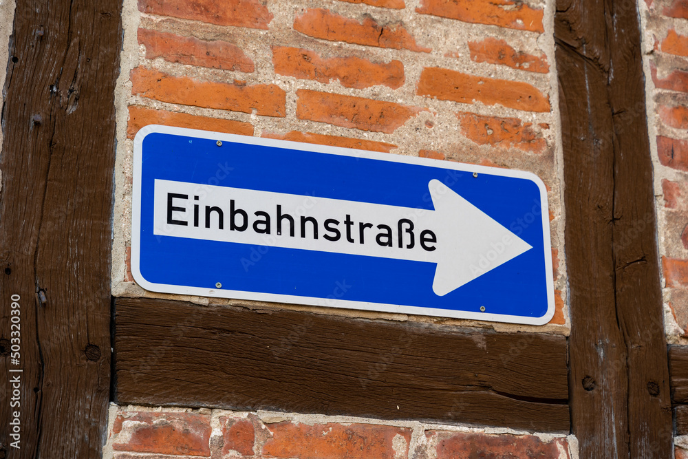 Obraz premium One way street traffic sign on a house wall in German language. Road sign of a white arrow on a blue metal plate. 'Einbahnstraße' is printed with black letters on the sign. Symbol for road users.