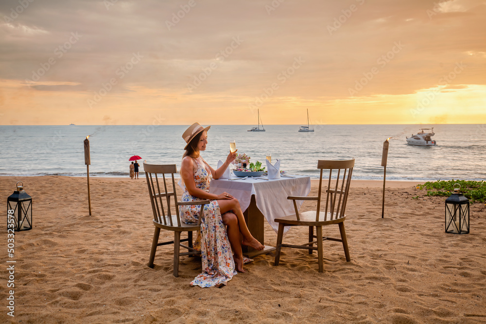 Romantic date on the sunset beach. Woman sitting alone on the table with gourmet food, drinking champagne from glass. Happy woman in luxury outdoor restaurant happy and smiling after proposal