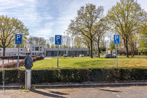 Public parking lot with spaces for handicap and charging station for electric cars, blue signs with a P, a drawing of a wheelchair and a car with an electric plug, building and cars in the background
