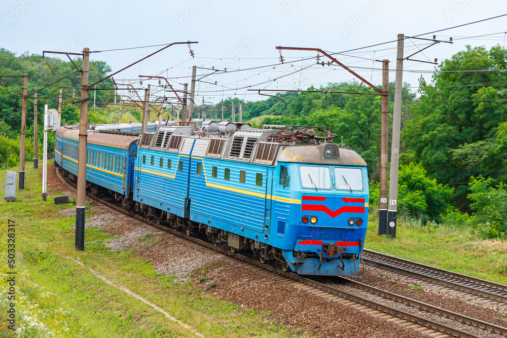 A powerful two-section blue locomotive pulls a long passenger train to the railway station. Trees with green leaves. Evening summer lighting.