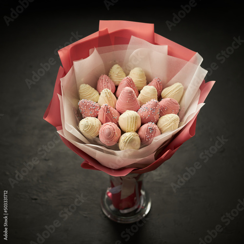 A bouquet of ripe strawberries covered with white and pink chocolate stands in a vase on a black background