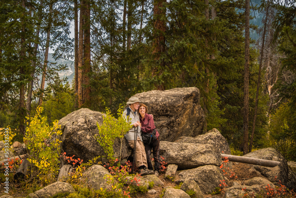 Environmental portrait of senior couple hiking in Rocky Mountains, Colorado, in autumn; rocks, pine trees and distant mountain in background