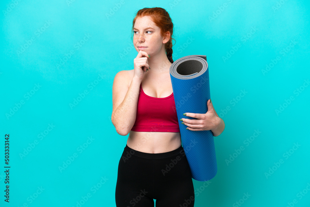 Young sport reddish woman going to yoga classes while holding a mat isolated on blue background having doubts and thinking Young sport reddish woman going to yoga classes while holding a mat isolated on blue background having doubts and thinking