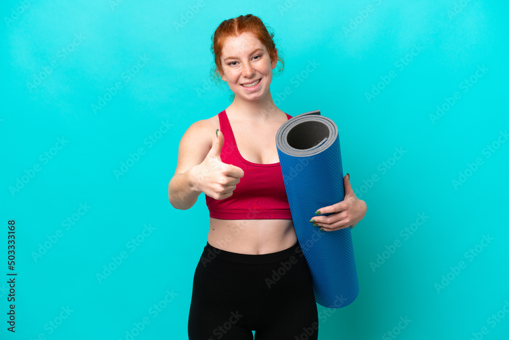 Young sport reddish woman going to yoga classes while holding a mat isolated on blue background with thumbs up because something good has happened Young sport reddish woman going to yoga classes while holding a mat isolated on blue background with thumbs up because something good has happened