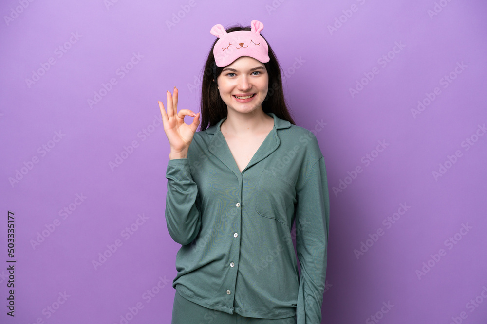 Young Russian woman in pajamas isolated on purple background showing ok sign with fingers