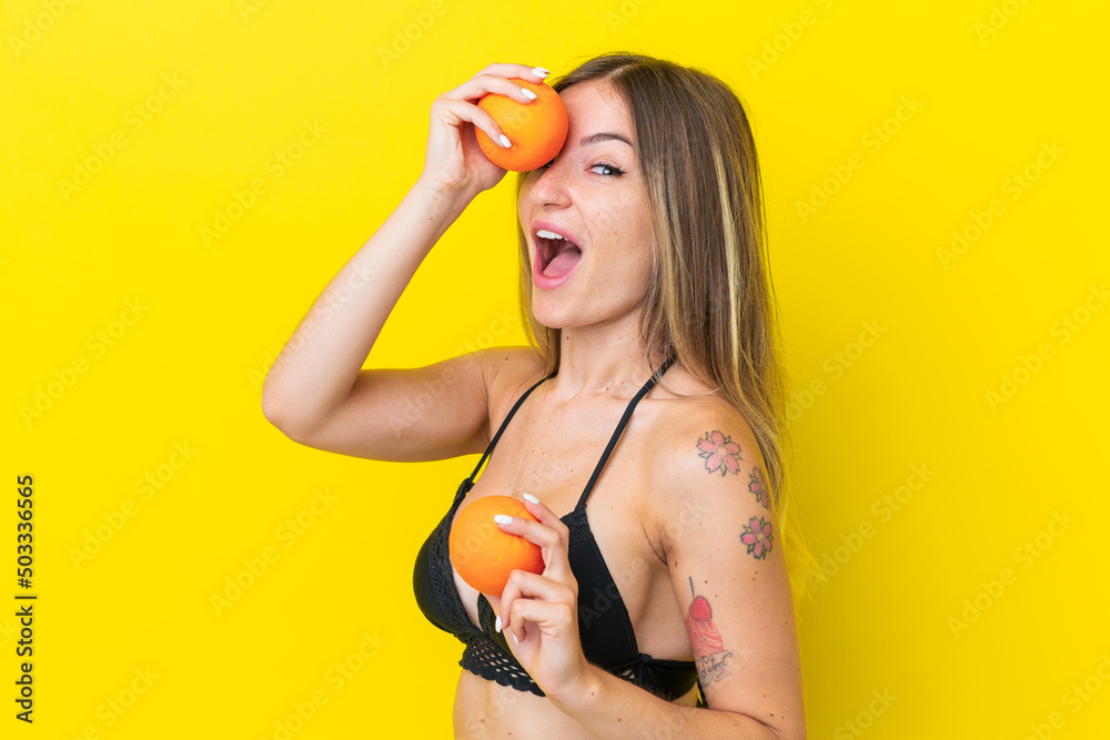 Young Romanian woman isolated on yellow background in swimsuit and holding an orange Young Romanian woman isolated on yellow background in swimsuit and holding an orange