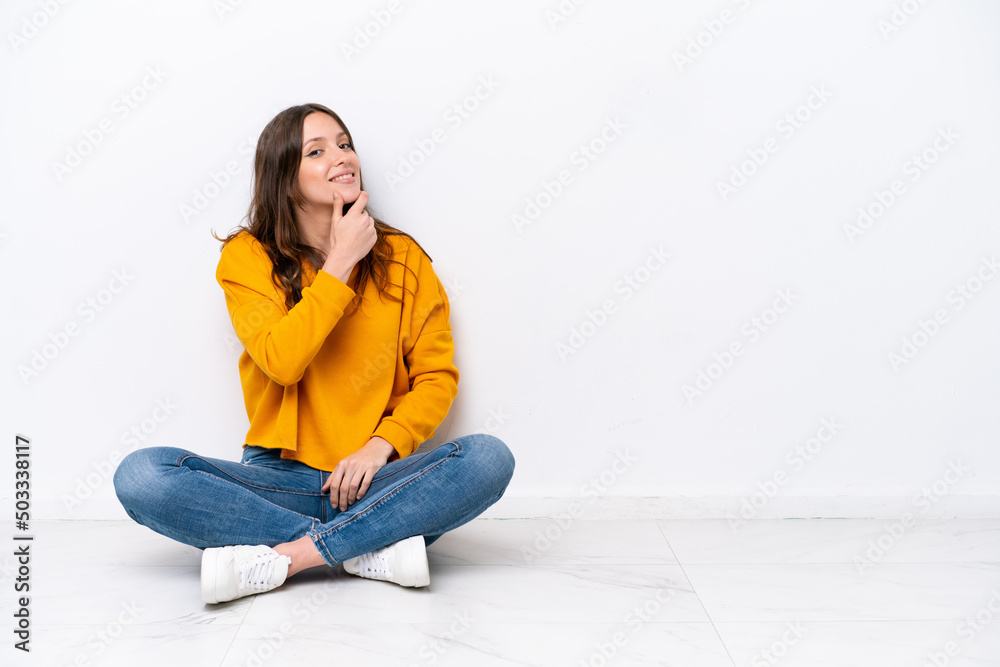 Young caucasian woman sitting on the floor isolated on white wall smiling