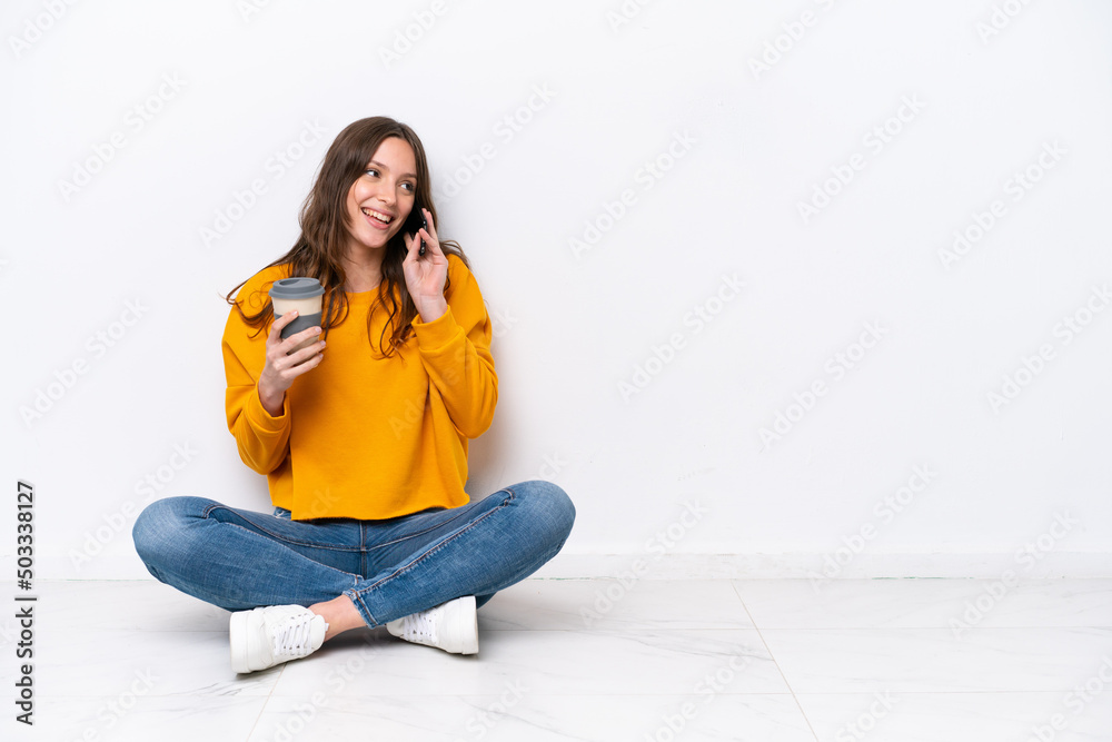 Young caucasian woman sitting on the floor isolated on white wall holding coffee to take away and a mobile Young caucasian woman sitting on the floor isolated on white wall holding coffee to take away and a mobile