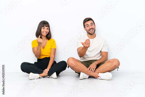 Young couple sitting on the floor isolated on white background inviting to come with hand. Happy that you came