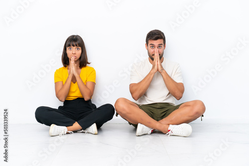 Young couple sitting on the floor isolated on white background keeps palm together. Person asks for something