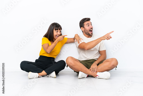 Young couple sitting on the floor isolated on white background presenting an idea while looking smiling towards