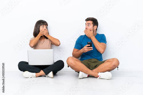 Young couple sitting on the floor holding pc and mobile phone isolated on white background covering mouth with hands for saying something inappropriate