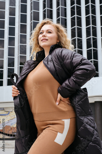 young blonde woman in trendy black quilted coat jacket oversized city walk advertising banner model plus size