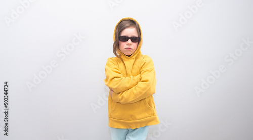 Child, Caucasian 6 years old, in a yellow sweatshirt with a hood and sunglasses on a gray background. Emotions on her face and crossed her arms on the gruli. © Fox