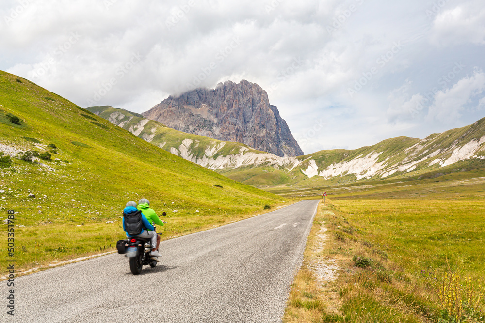 Two people riding a motorcycle on a mountain road. A couple of tourists on motorcycles on a road in the Apennines in Abruzzo, Italy. Campo Imperatore plateau with the Gran Sasso peak in the background