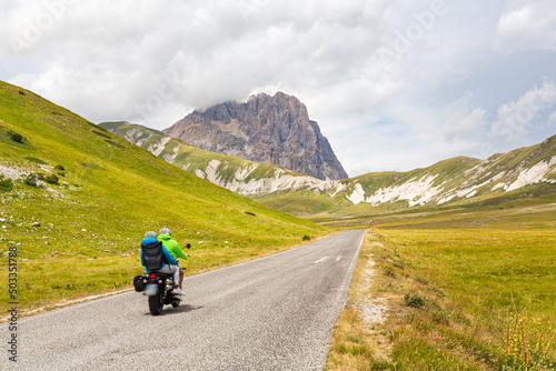 Two people riding a motorcycle on a mountain road. A couple of tourists on motorcycles on a road in the Apennines in Abruzzo  Italy. Campo Imperatore plateau with the Gran Sasso peak in the background