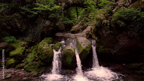 Beautiful Schiessentümpel waterfall under the arch of an old stone bridge in the woods of Mullerthal Luxembourg. Low angle view forward motion, low over the flowing water.