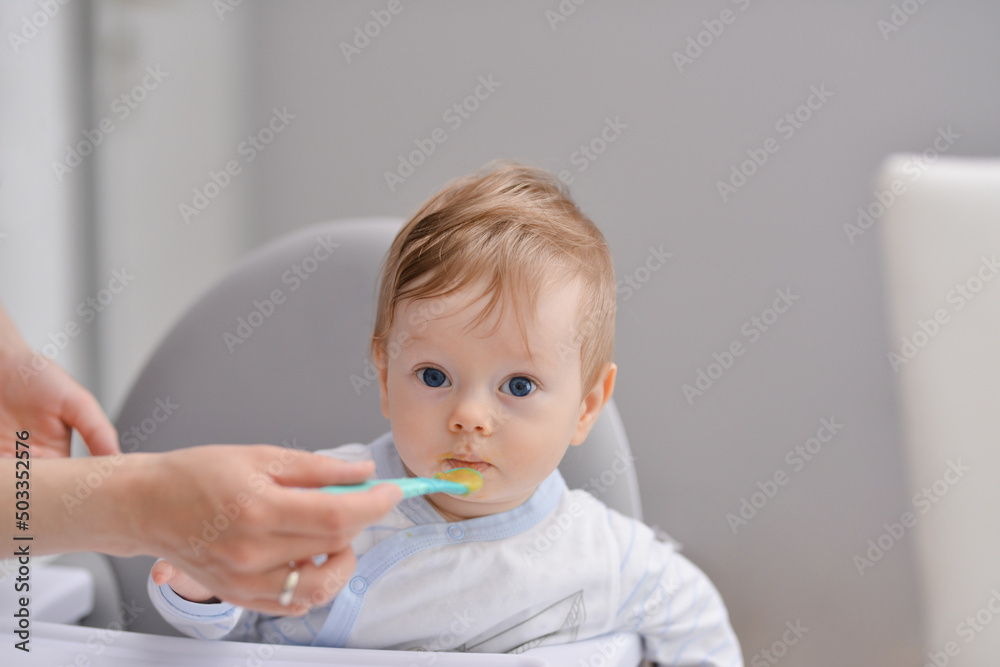 Mother feeding baby with spoon, baby boy child sitting eating food on chair at home Mother feeding baby with spoon, baby boy child sitting eating food on chair at home