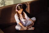 A brunette little girl in pink headphones sits on the couch, looks into a digital tablet.