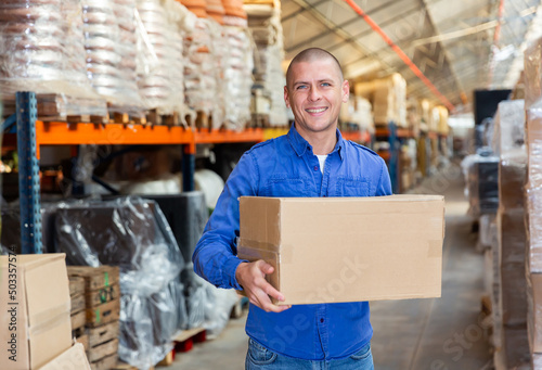 Positive warehouse worker standing, holding pasteboard box, looking at camera and smiling.