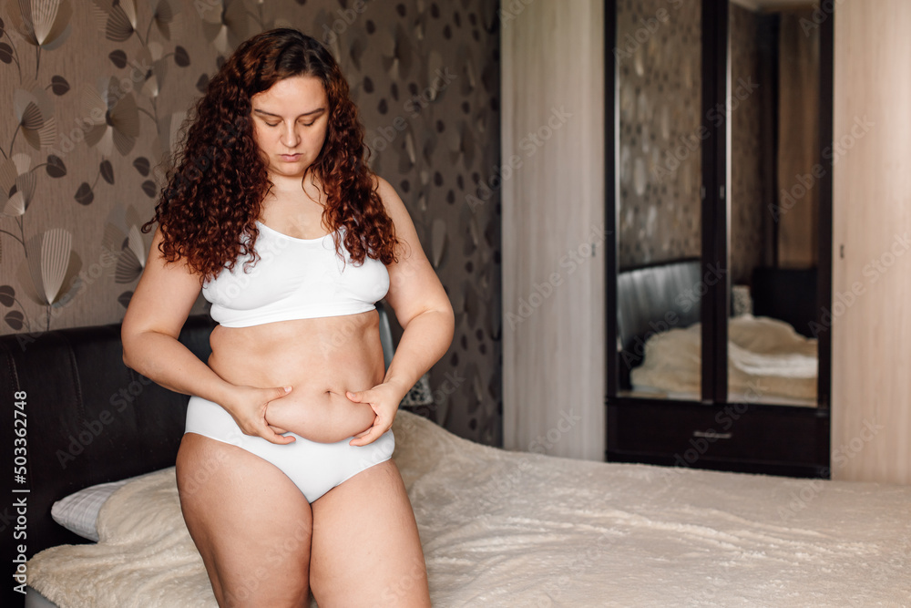 Curly plus size young woman in white underwear standing near bed in bedroom  and pinching folds on sagging stomach. Fat burning treatment of thick tummy.  Weight loss program, anticellulite massage. Stock Photo