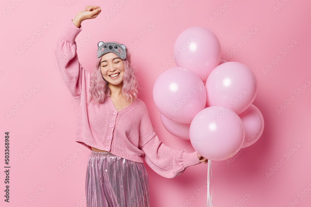 Positive cheerful Asian woman dances carefree keeps arm raised up holds bunch of inflated balloons wears casual jumper and pleated skirt sleepmask on forehead enjoys party time. Fesitve event