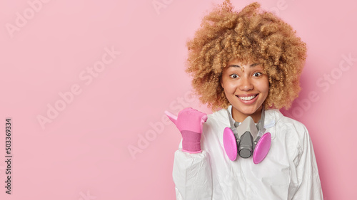Positive woman in white protective costume uses gas mask points away on blank space against pink background tells about radiation hazard protects herself from environmental pollution. Look there